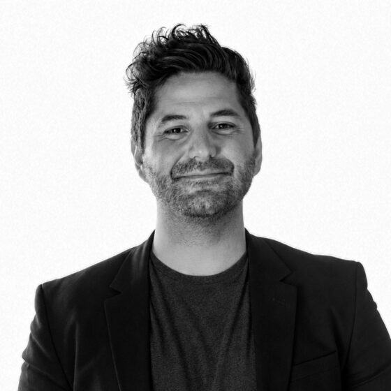 Head-and-shoulders portrait of a smiling man with dark hair and stubble, wearing a blazer and t-shirt against a light background.